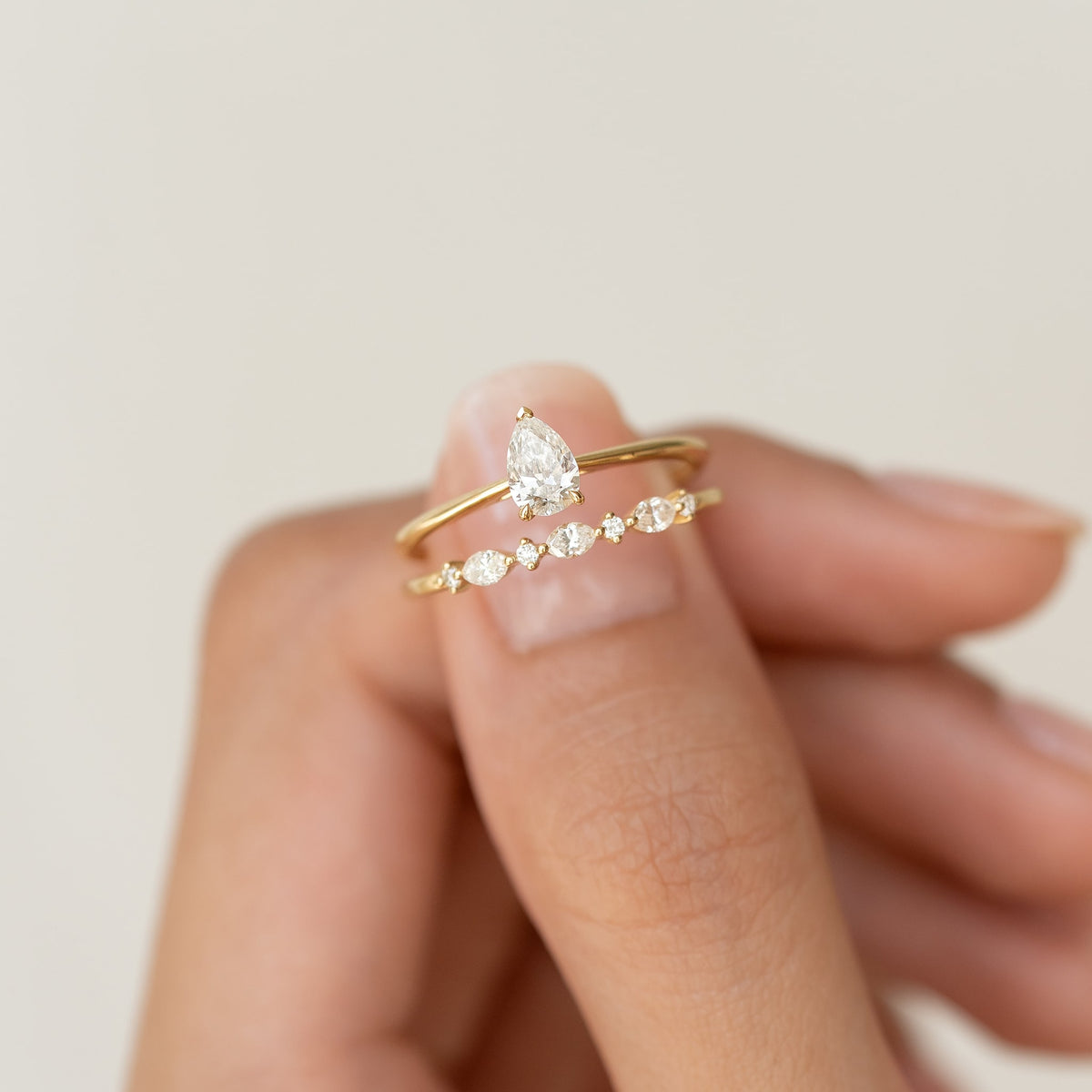 Close-up of a hand holding two delicate Gold rings with diamonds. One features a pear-shaped diamond, the other showcases multiple round diamonds.