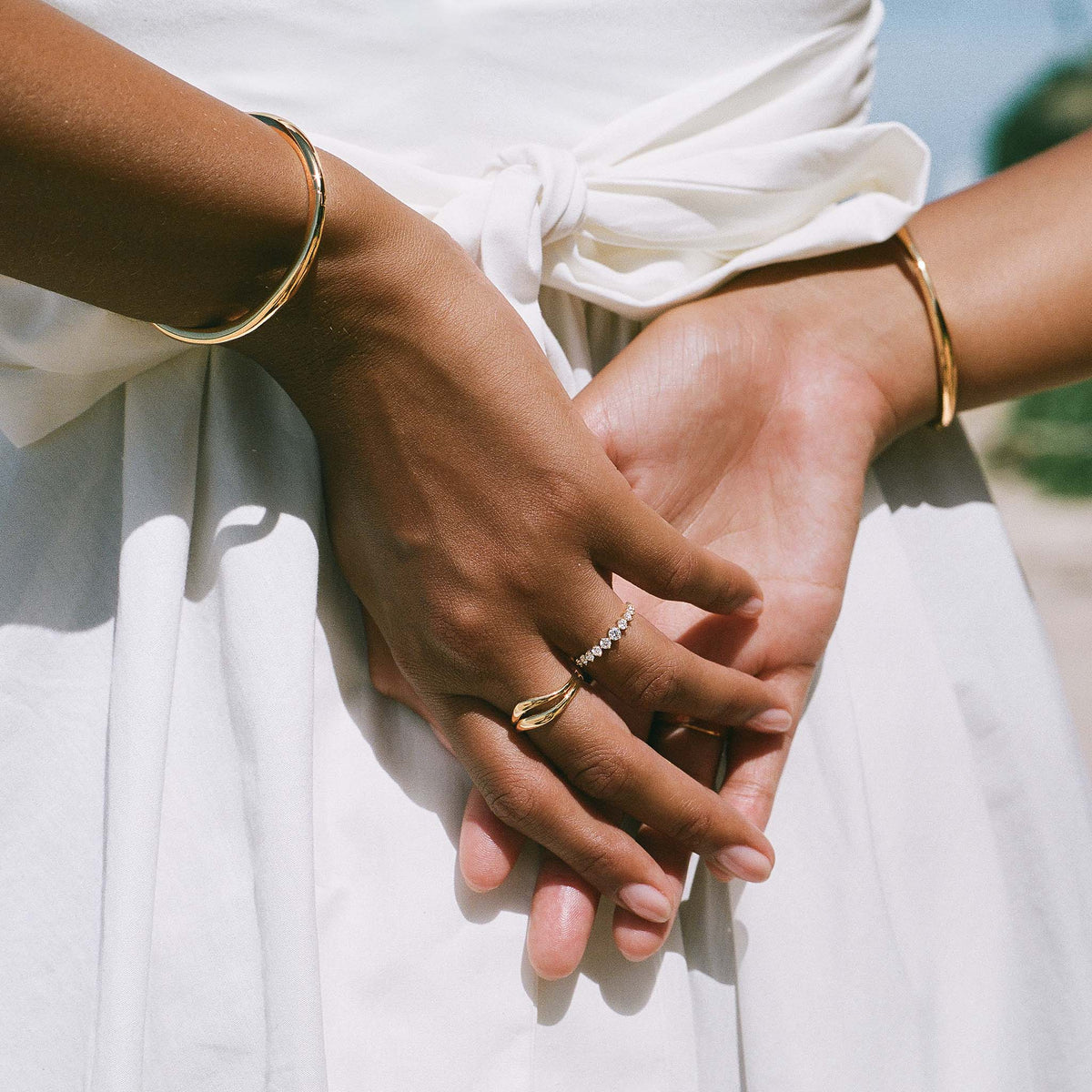 Close-up of hands with gold bangles and the Organic Wavy Ring - Nyssa worn on a finger over a white dress
