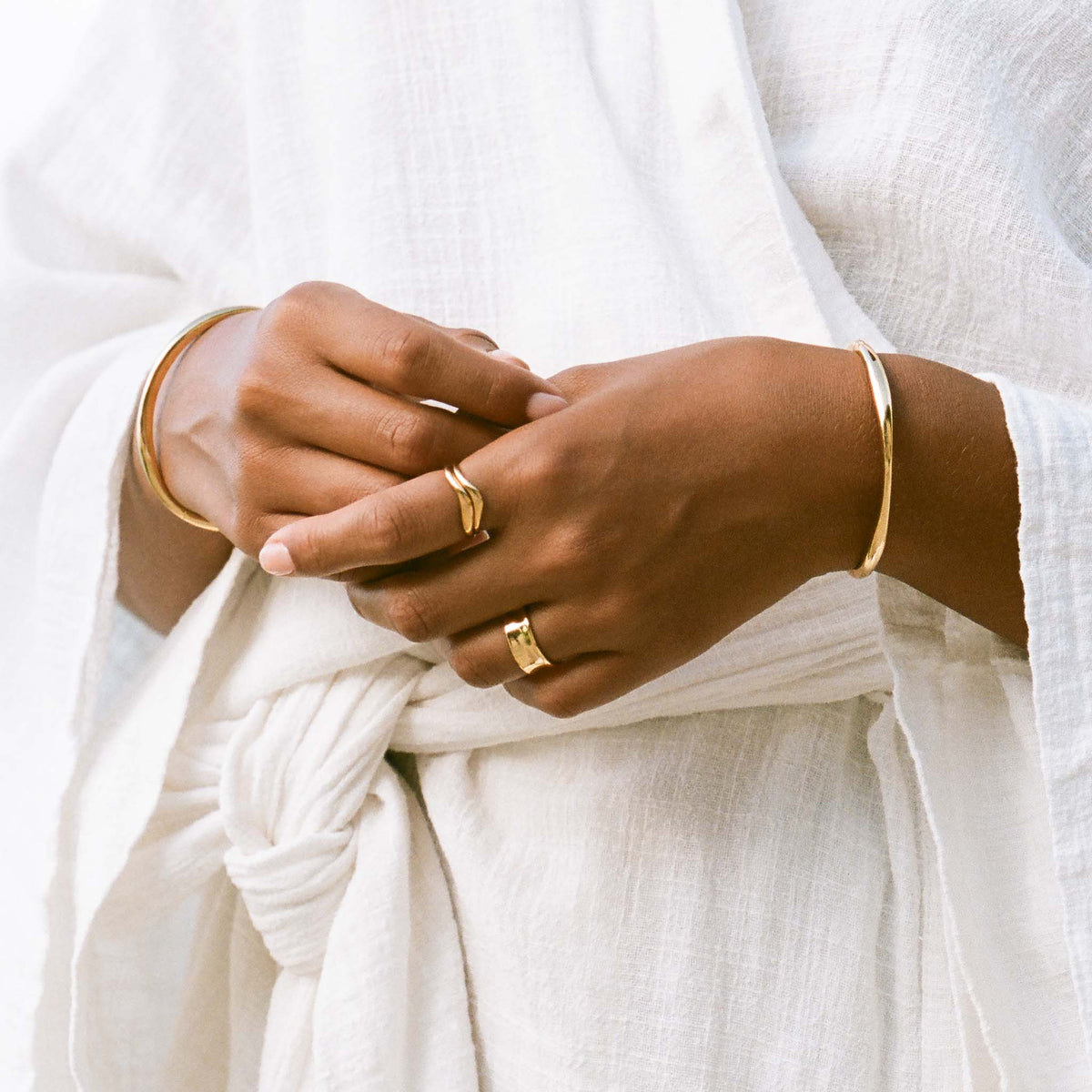 Close-up of a person wearing the Gold Organic Bangle S - Aelia with gold rings and a white textured garment background