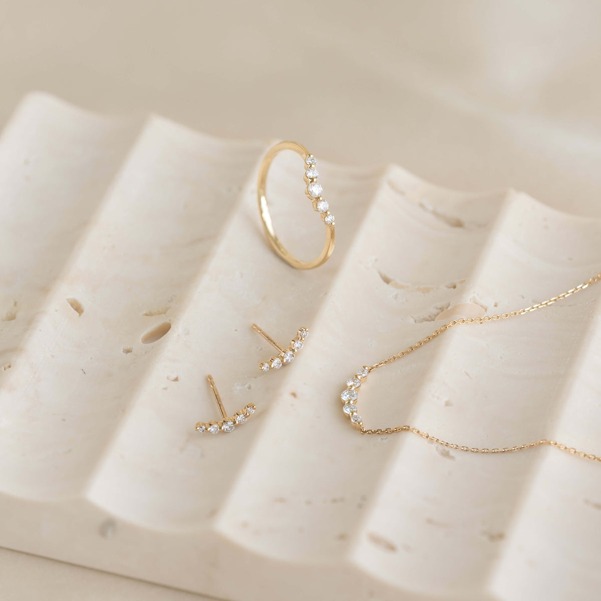 Close-up of Diamond Curved Bar Necklace 14k Gold - Elodie with matching curved bar earrings and ring on beige stone surface