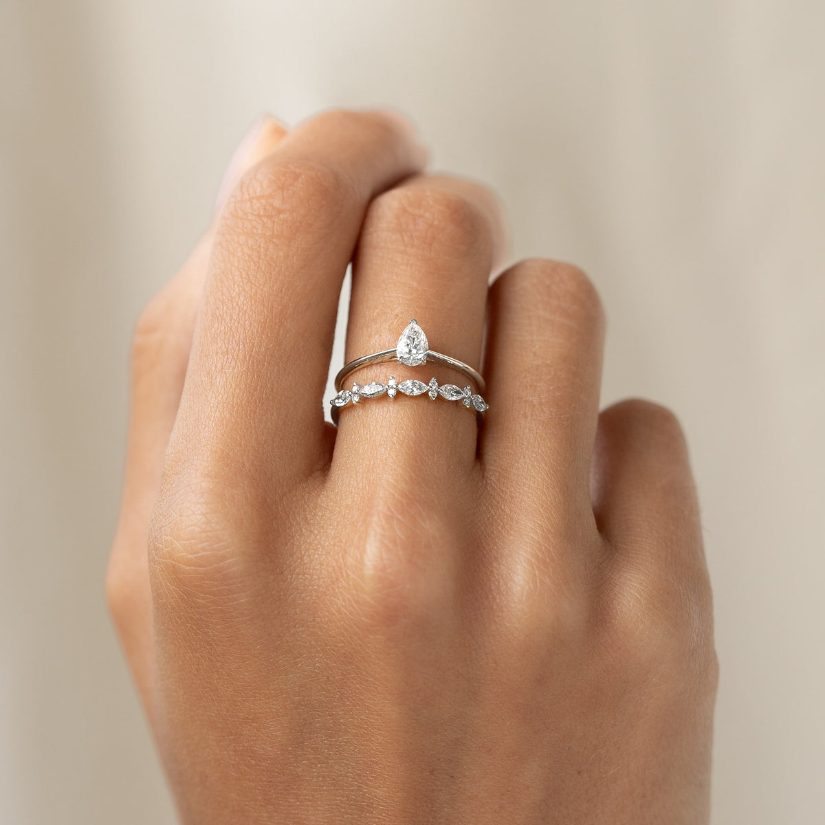 A hand wearing two elegant White Gold  rings: a pear-shaped diamond solitaire ring and a band with small marquise and round diamonds, on a light background.
