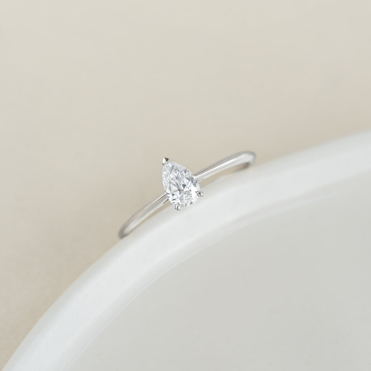 A delicate White Gold ring features a single pear-shaped diamond on a smooth, minimalist band, set against a soft, neutral background, conveying elegance and simplicity.