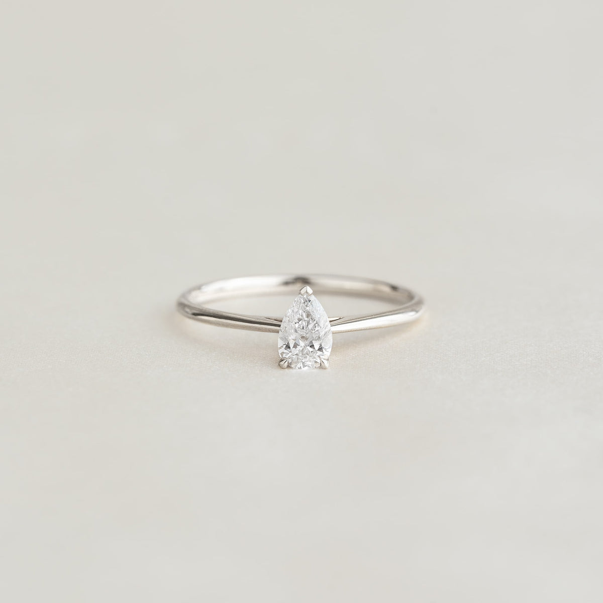 A minimalist White Gold ring with a pear-shaped diamond set on a thin, elegant band sits against a soft, neutral background, evoking simplicity and elegance.