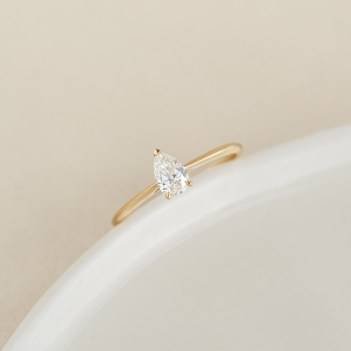 A delicate Gold ring features a pear-shaped diamond set elegantly in the center, positioned on a smooth, light surface, embodying simplicity and elegance.
