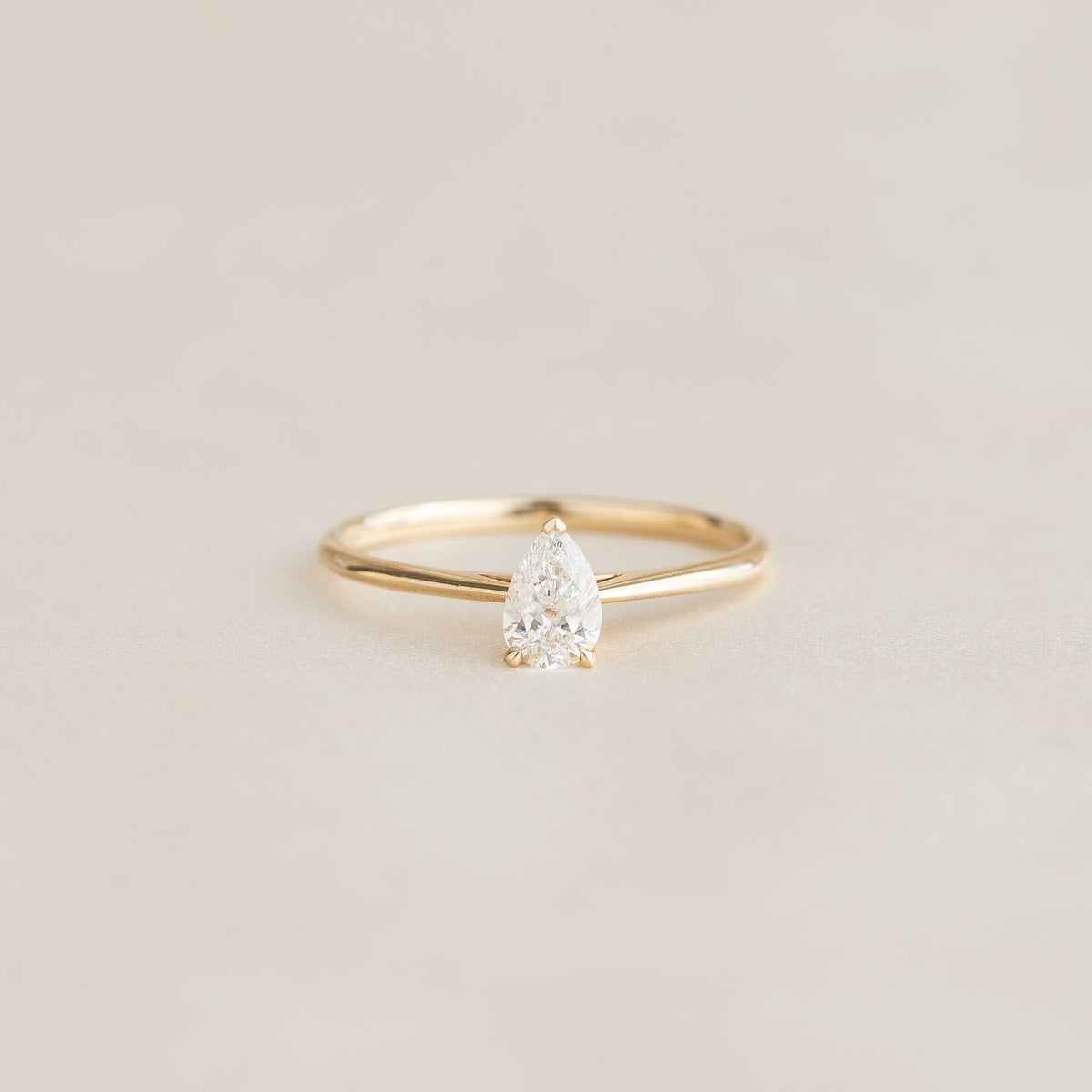 A delicate Gold ring with a pear-shaped diamond set on a soft beige background, exuding elegance and simplicity.