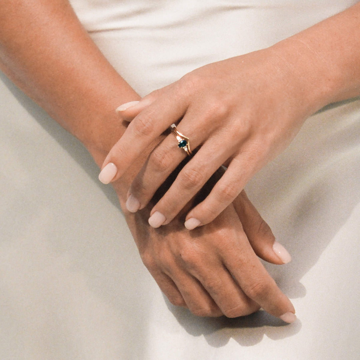 A top-down photograph shows a person's hands gently crossed over each other against a soft, light-colored fabric background, showcasing a Yellow Gold ring stack.