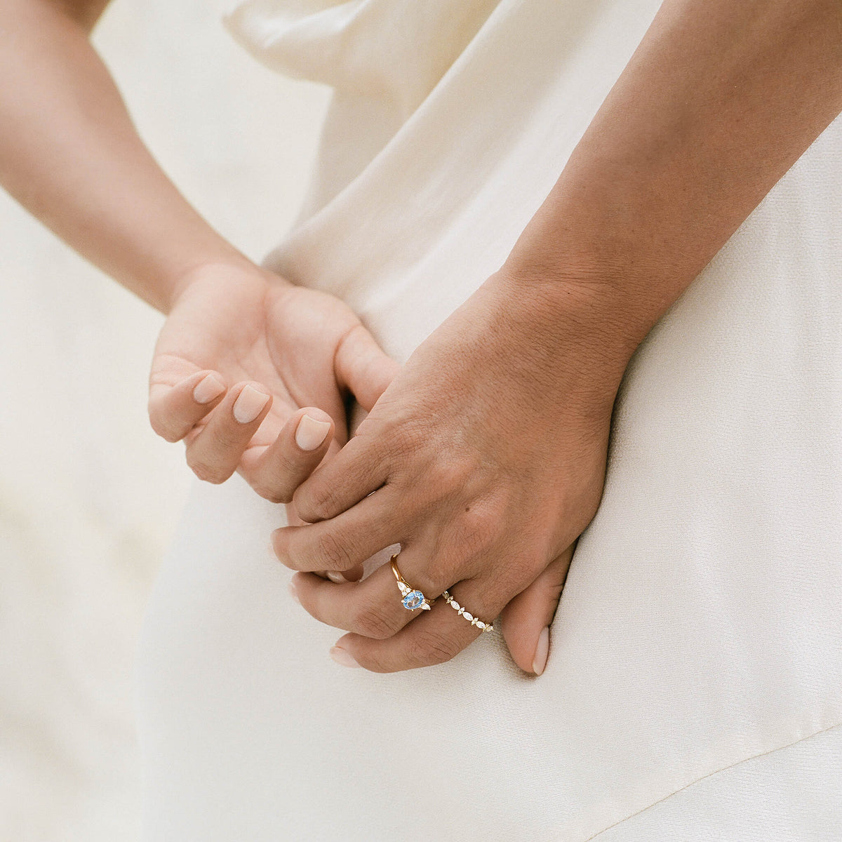 A close-up photograph shows a person's hands gently clasped behind her back, showcasing a fine Yellow Gold jewelry pairing on her fingers.