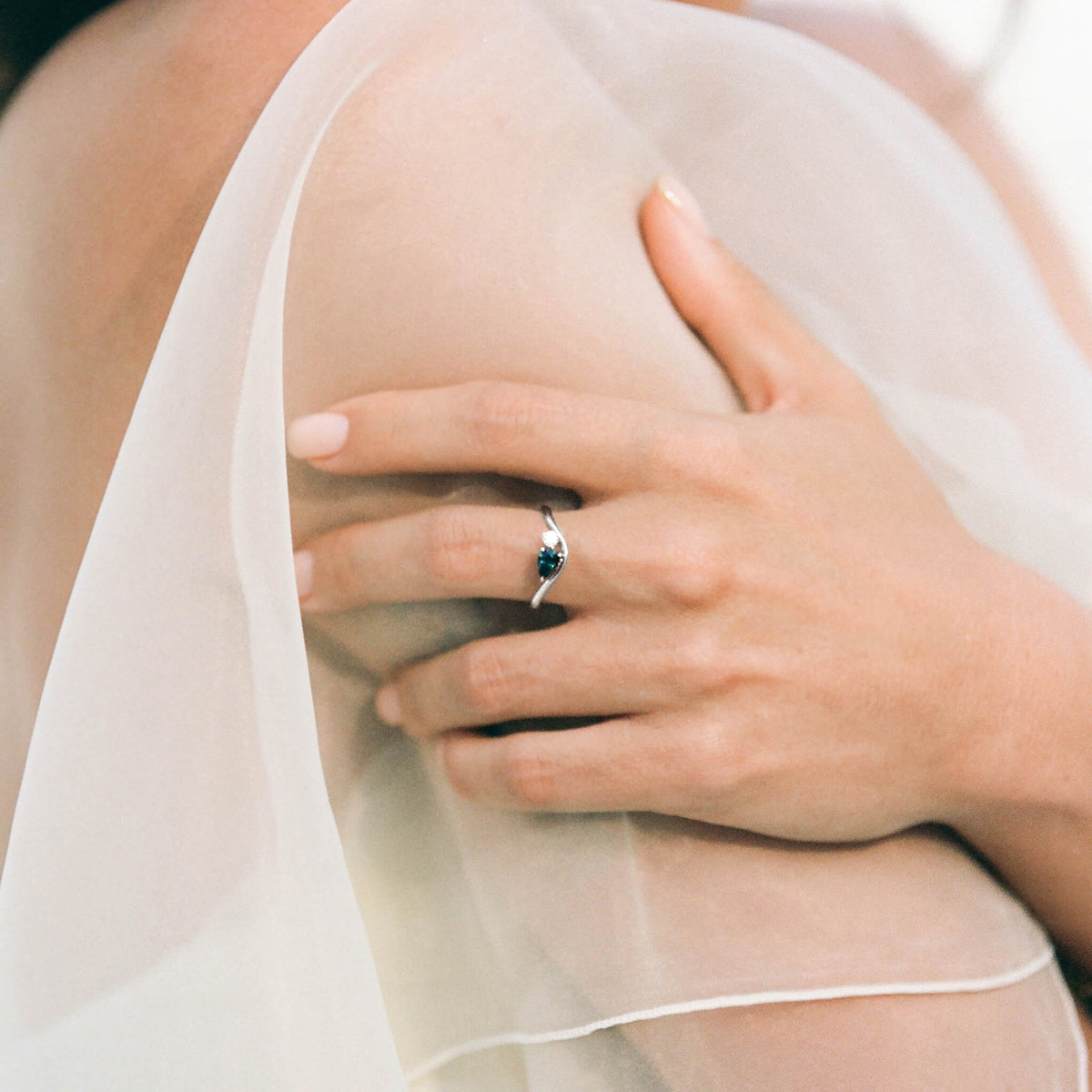 The individual is wearing an elegant White Gold ring on their finger, which features a pear-cut, deep blue sapphire accented by a small white diamond.