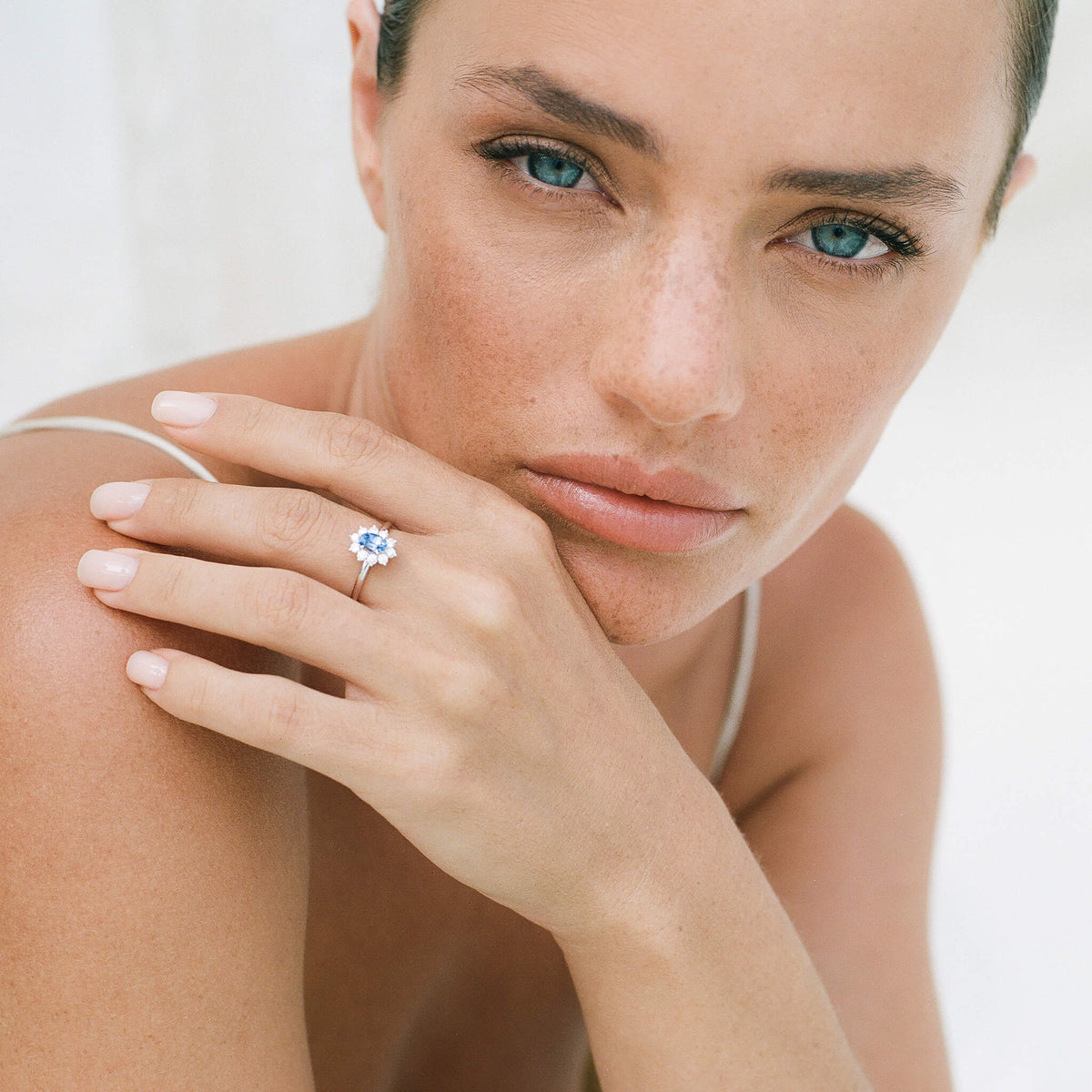 Her hand rests gently against her chin and shoulder, showcasing the White Gold Sarai sapphire halo ring worn on her middle finger.