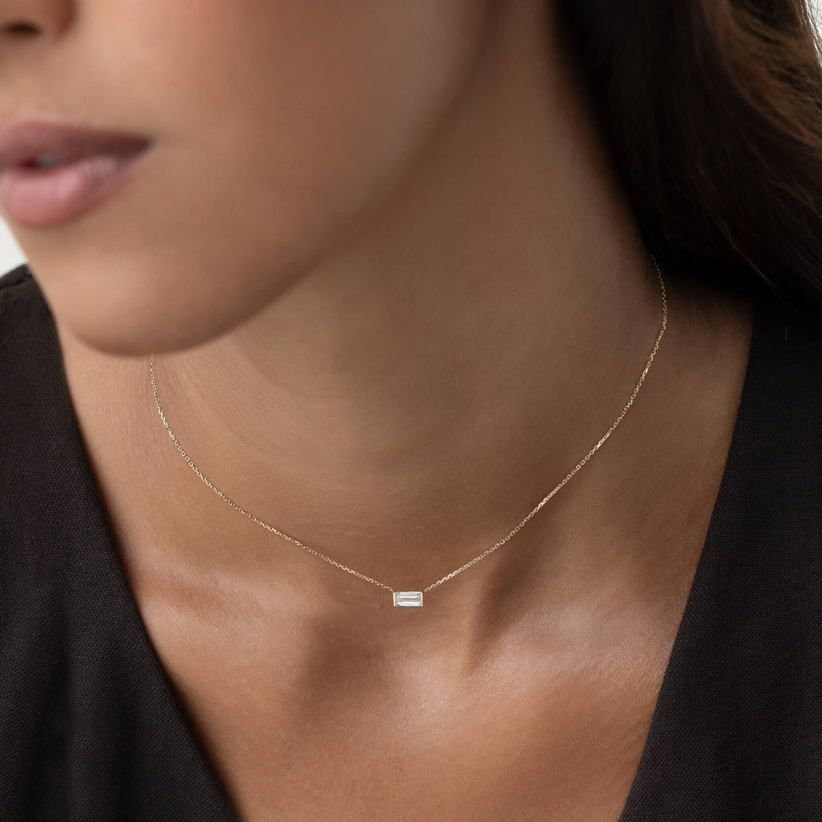 Close-up of a woman wearing a delicate Gold chain necklace with a small rectangular clear gemstone pendant, paired with a black V-neck top.