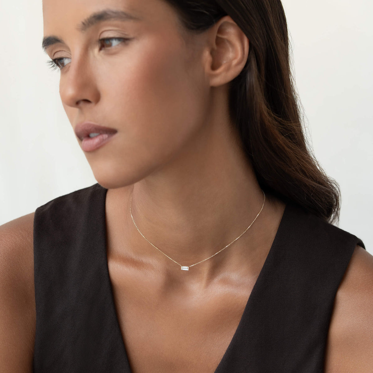 Woman wearing a delicate Gold chain necklace with a small rectangular clear gemstone pendant, styled with a black sleeveless V-neck top against a soft white background.