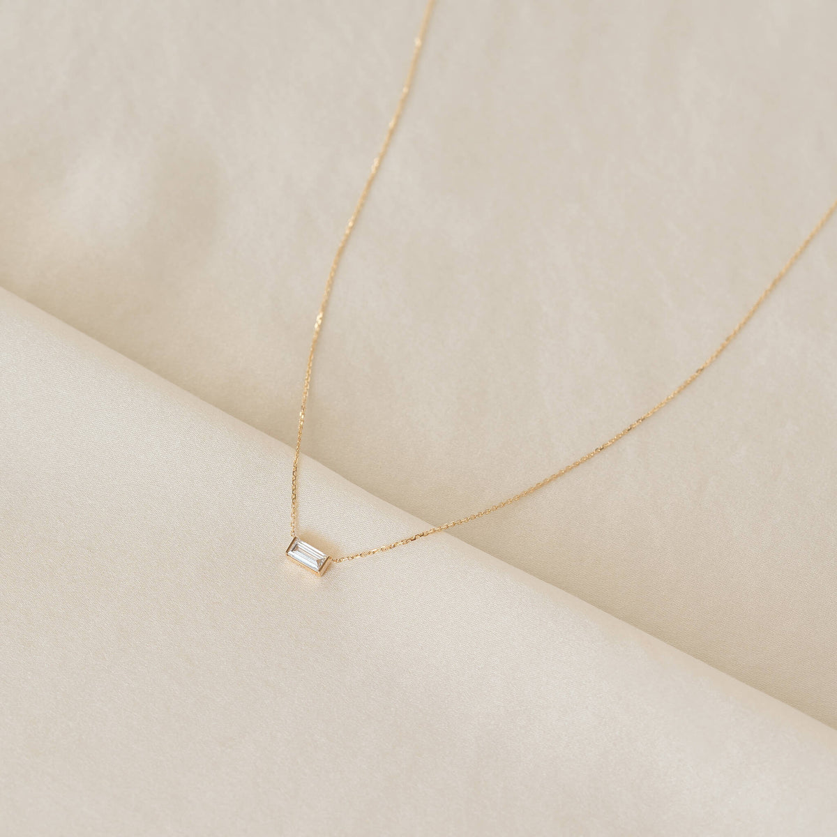 Delicate Gold chain necklace with a small rectangular baguette-cut clear gemstone pendant, arranged on soft cream fabric with gentle folds.