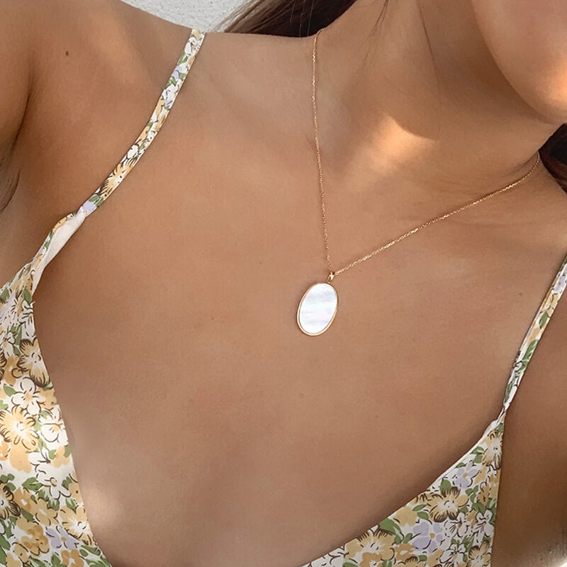 Close-up of a woman wearing a delicate gold necklace with an oval white pendant and a floral dress with thin straps