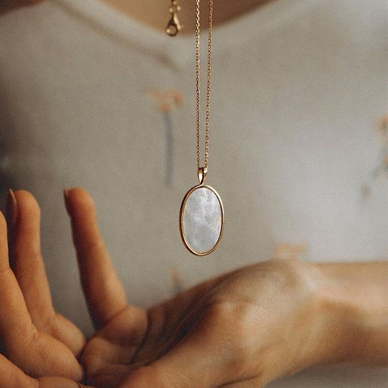 Close-up of a hand reaching toward a delicate gold chain necklace with an oval white pendant against a soft background
