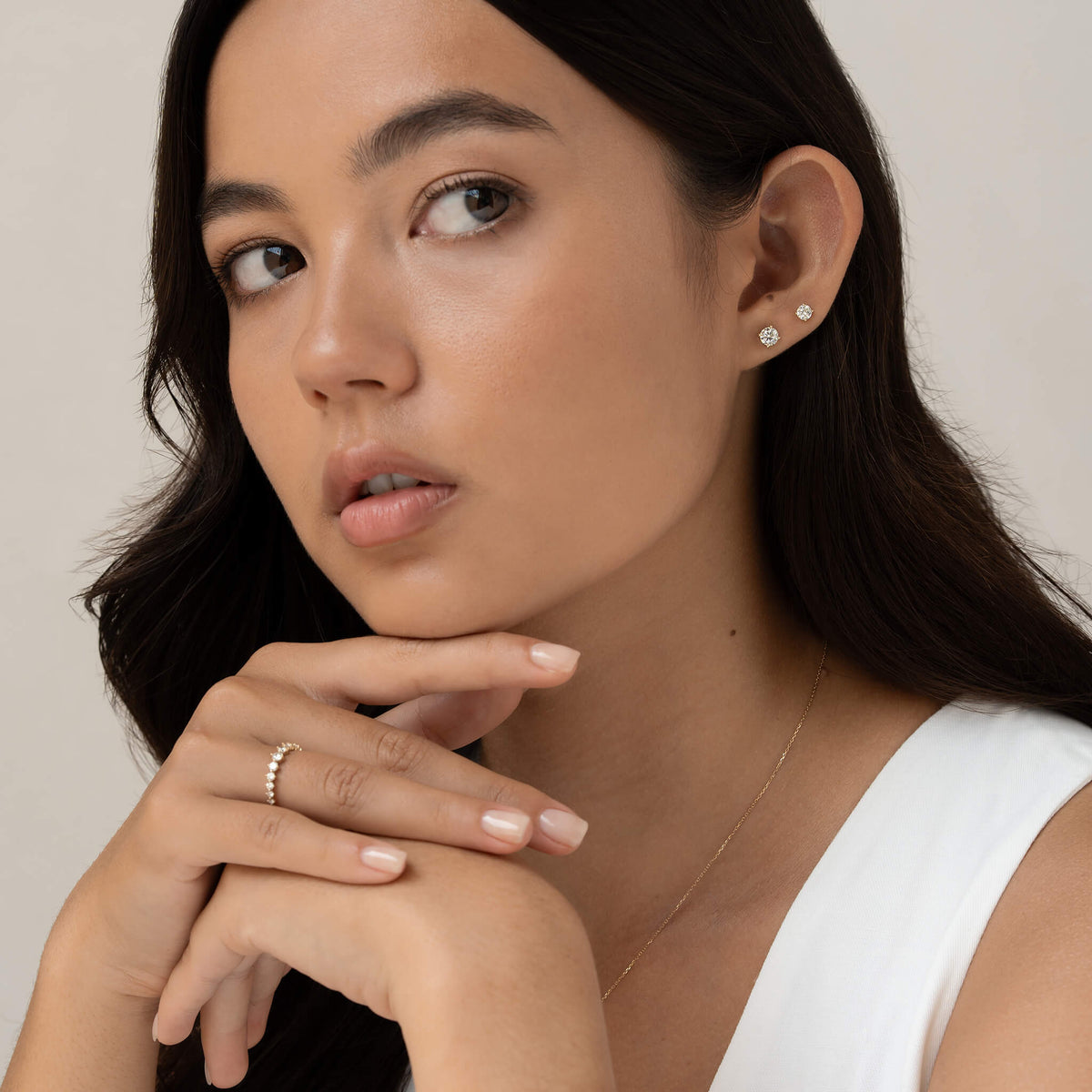 A woman gazes thoughtfully, resting her chin on her hand. She wears delicate stud earrings and a thin ring. Her expression is calm and contemplative.