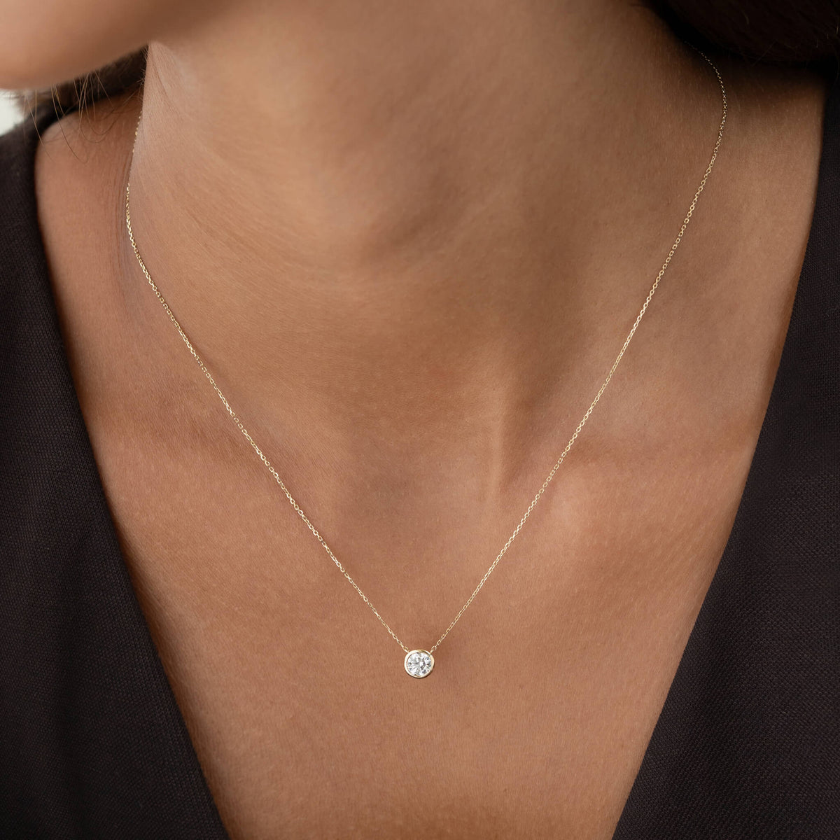 A close-up view of a person wearing a delicate Gold chain necklace with a single, round diamond solitaire pendant held in a minimalist bezel setting, worn against a simple dark-colored top.