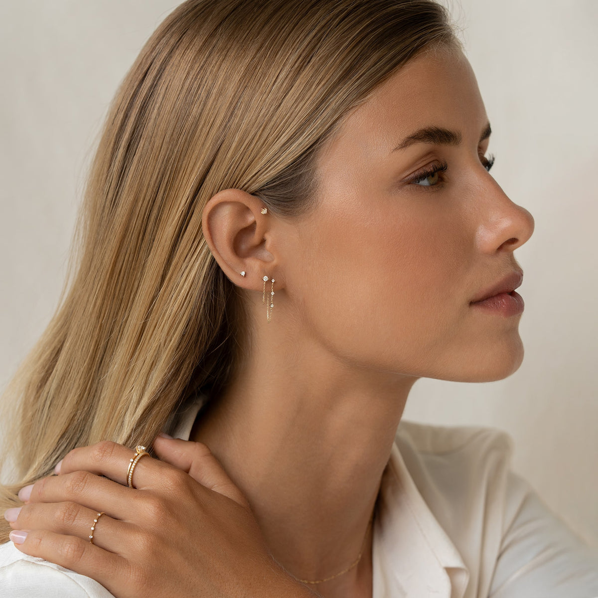 A woman with blonde hair in a side profile wears multiple Gold earrings and rings, looking serene. She wears a white shirt, enhancing a minimalist and elegant tone.