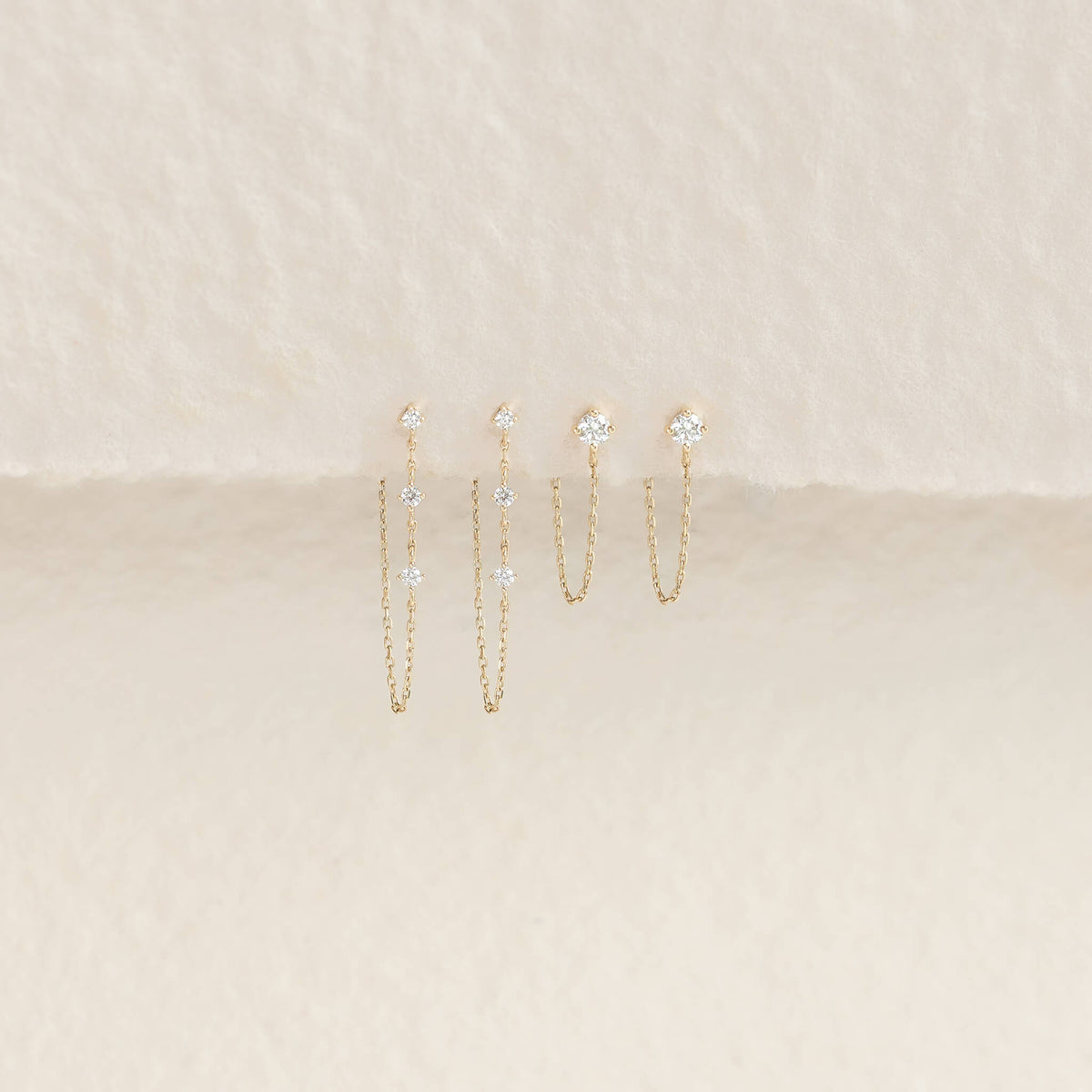 Elegant Gold earrings with hanging chains and sparkling gems on a textured beige background, exuding a delicate and luxurious feel.