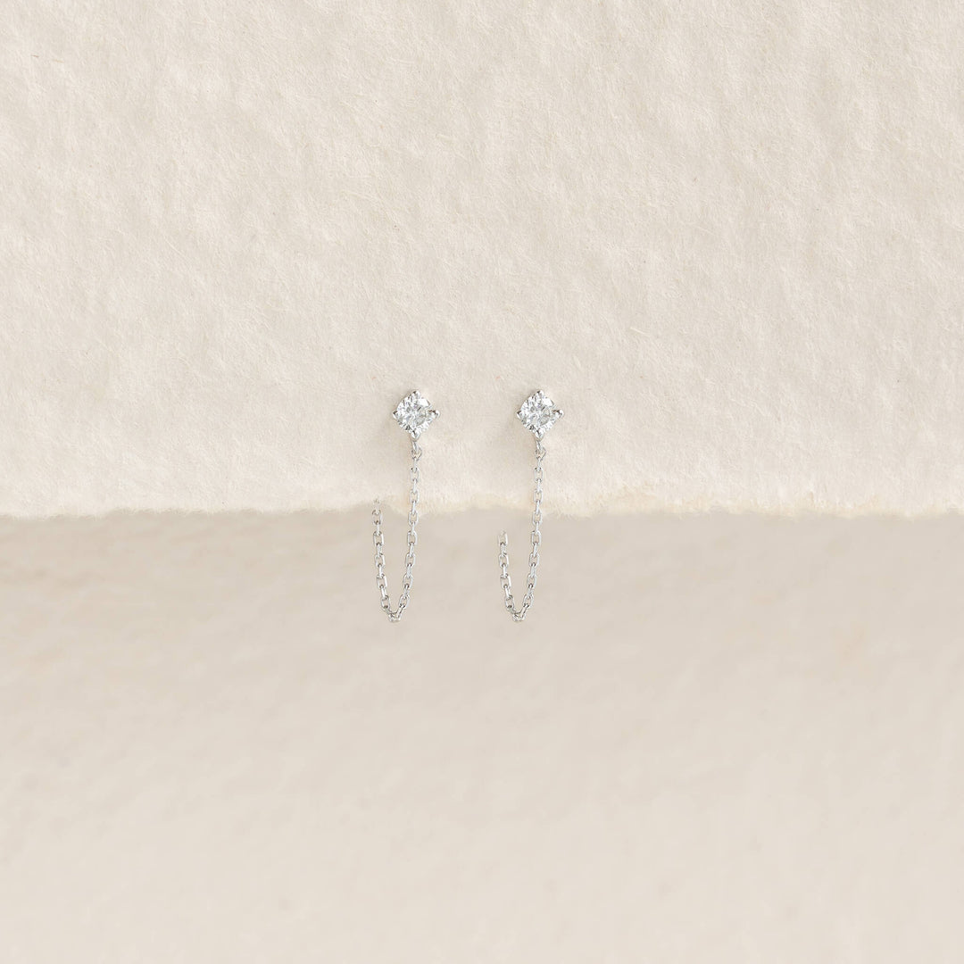 Delicate White Gold with small diamond studs and thin chains, elegantly displayed on a textured, cream-colored background. Elegant and minimalist.