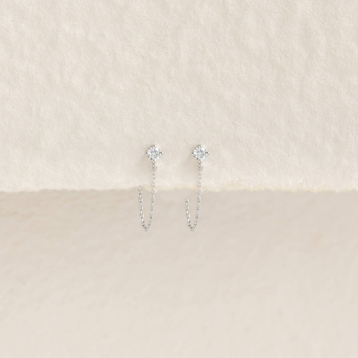 Delicate White Gold with small diamond studs and thin chains, elegantly displayed on a textured, cream-colored background. Elegant and minimalist.