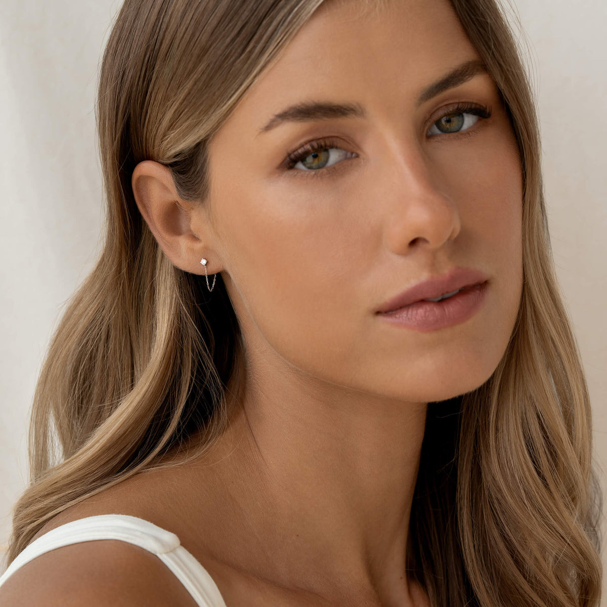 Woman with long blonde hair and soft expression gazes at the camera. She wears a White Gold small hoop earring and a white top.