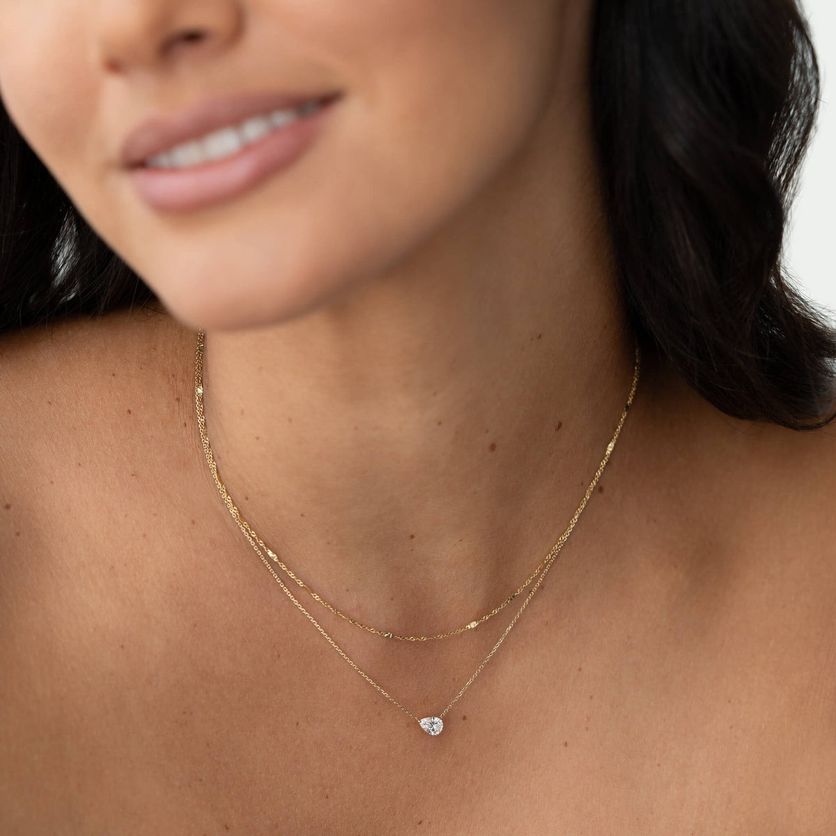 Close-up of a woman wearing layered Gold necklaces, including a smooth chain and a delicate pear-shaped gemstone pendant.