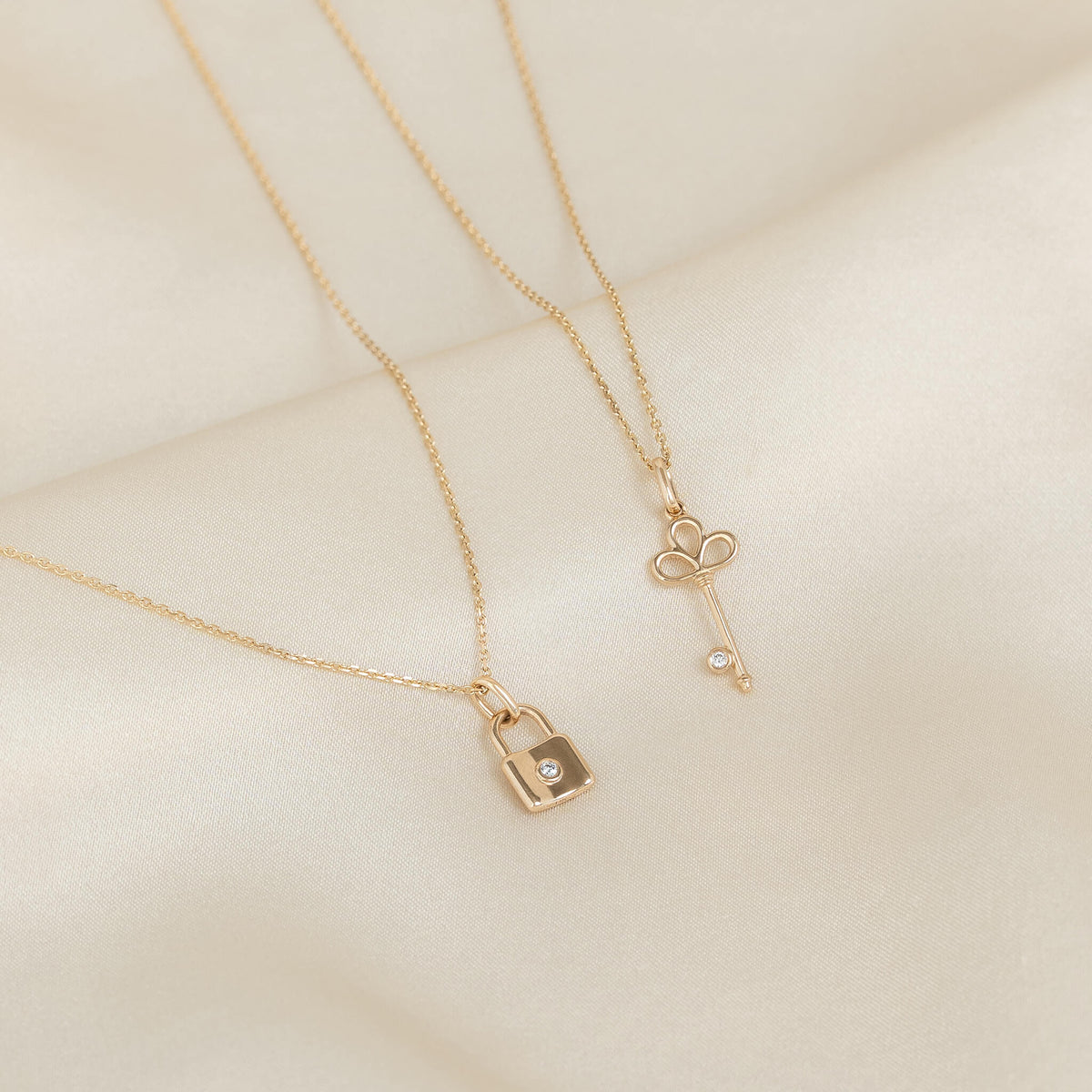 Two delicate Yellow Gold necklaces displayed on a soft textured surface, featuring padlock and key-shaped pendants with small diamond accents.