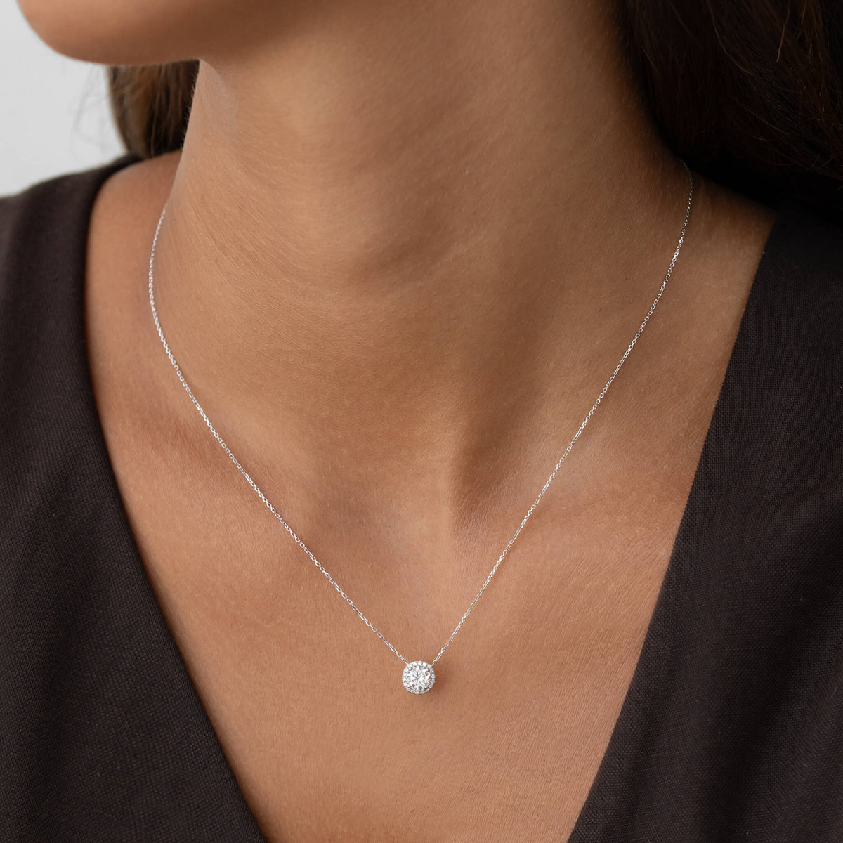 A close-up shot of a person wearing a delicate White Gold chain necklace with a small, round brilliant-cut diamond halo pendant, worn against a dark-colored V-neck top.