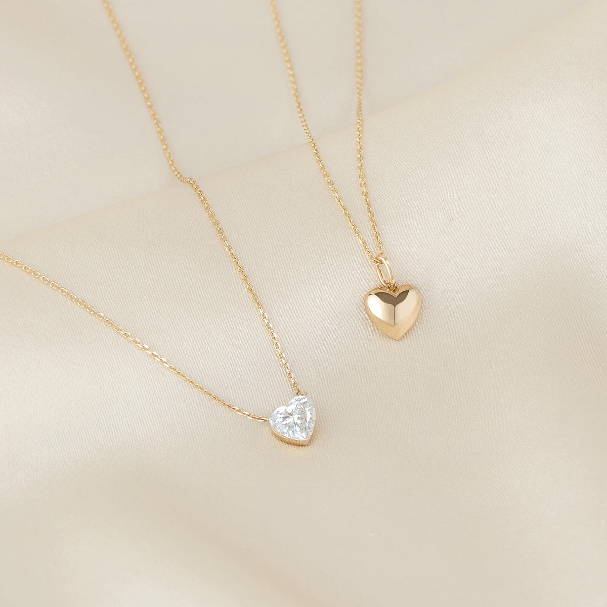 Two delicate Yellow Gold necklaces displayed together, one featuring a polished heart-shaped pendant and the other a sparkling heart-shaped gemstone pendant, on a soft neutral fabric background.