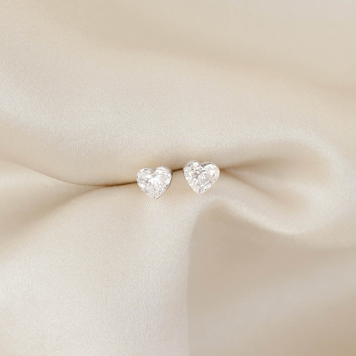 Pair of White Gold heart-shaped diamond stud earrings resting on softly draped, cream-colored fabric, highlighting their sparkle against a smooth, neutral background.