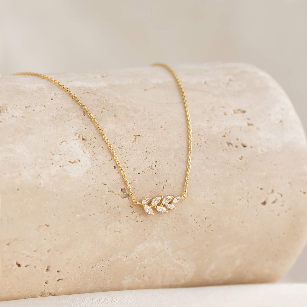 Gold necklace with a delicate leaf pendant adorned with small diamonds, elegantly displayed on a textured beige stone. The tone is elegant and refined.