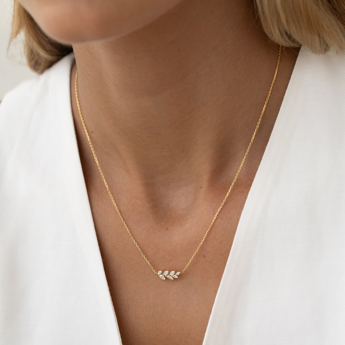 A woman's neck wearing a delicate Gold necklace with a small leaf pendant, over a white blouse. The mood is elegant and minimalist.