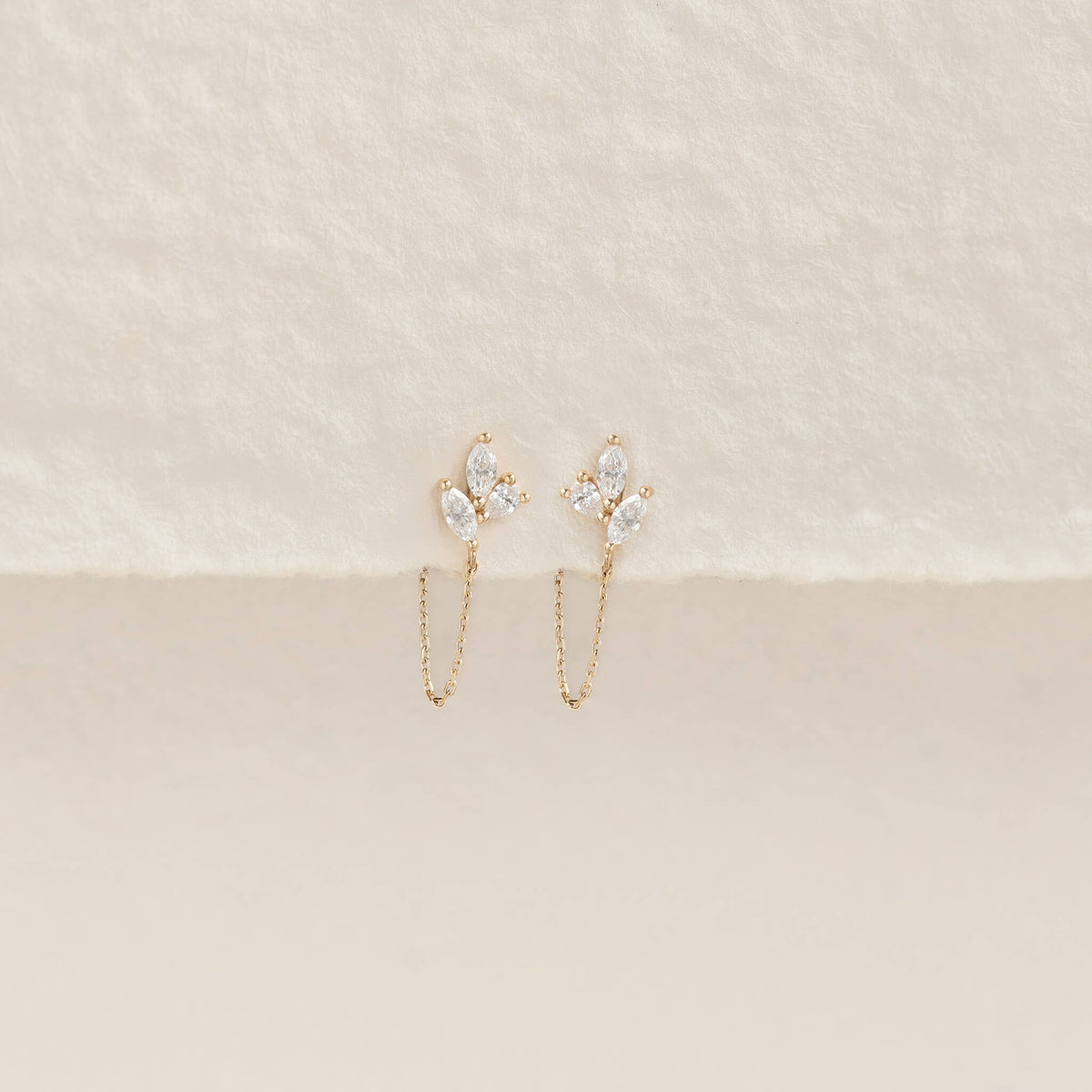 Elegant earrings feature a delicate Gold chain and leafy cluster of sparkling white gems, set against a soft beige textured background.