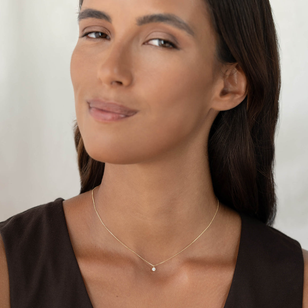 A close-up portrait of a woman with dark hair wearing a minimalist, delicate Gold chain necklace with a tiny round brilliant diamond pendant, styled with a simple dark brown top.