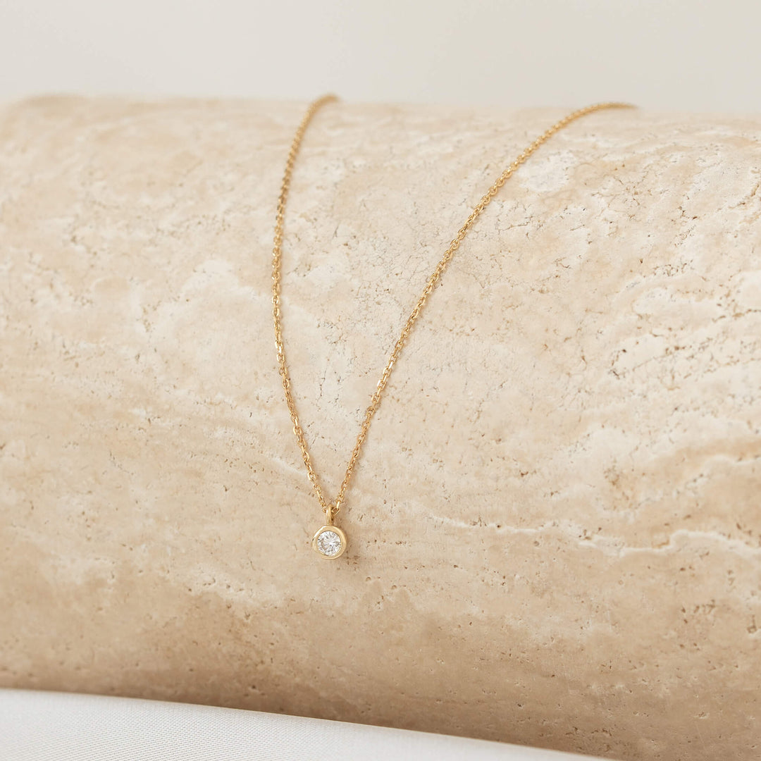 A close-up shot of a delicate Gold necklace with a small, round diamond solitaire pendant in a bezel setting, draped over a textured, light beige stone surface.