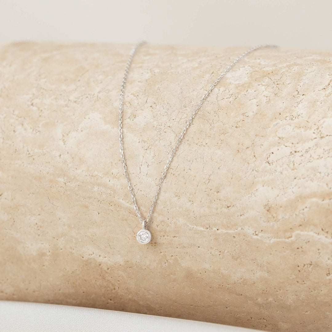 A close-up shot of a delicate White Gold necklace with a small, round diamond solitaire pendant in a bezel setting, draped over a textured, light beige stone surface.