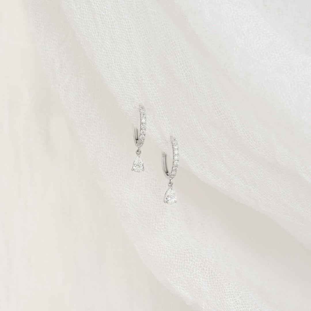A pair of white gold hoop earrings with small diamonds and teardrop-shaped diamond dangles displayed on a white textured fabric background.