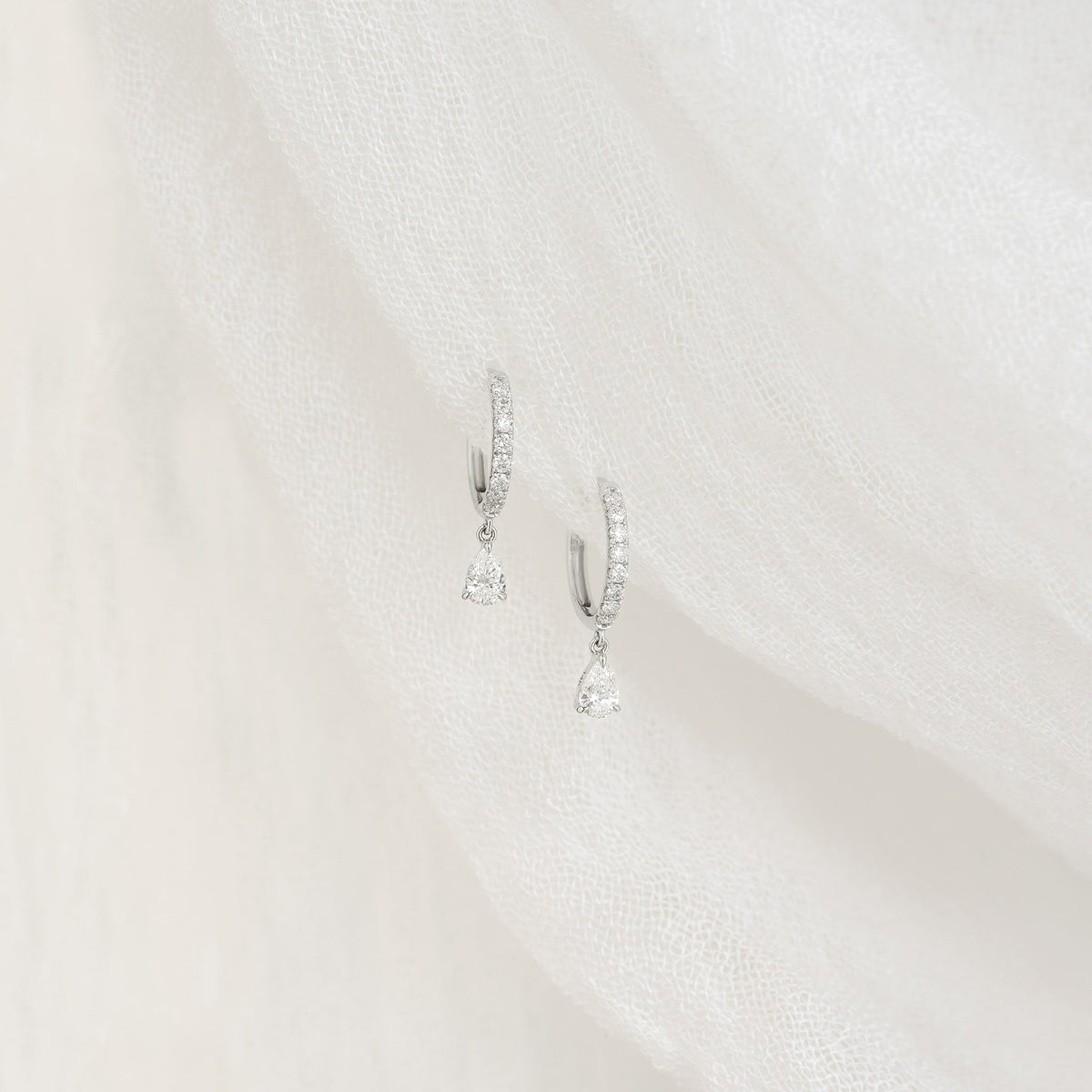 A pair of white gold hoop earrings with small diamonds and teardrop-shaped diamond dangles displayed on a white textured fabric background.