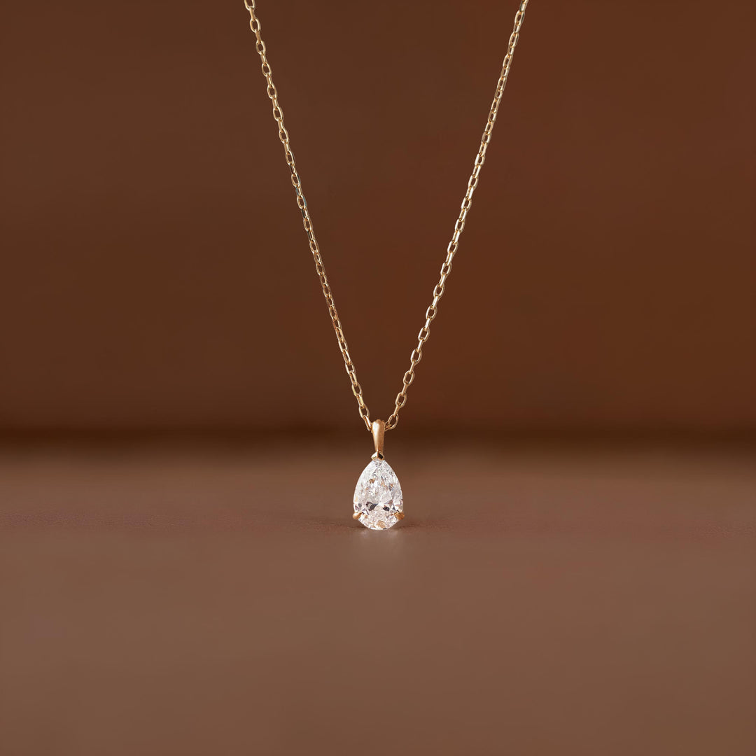 A close-up shot of a delicate Gold necklace featuring a brilliant, pear-shaped diamond pendant suspended from a fine link chain against a soft, neutral brown-colored background.
