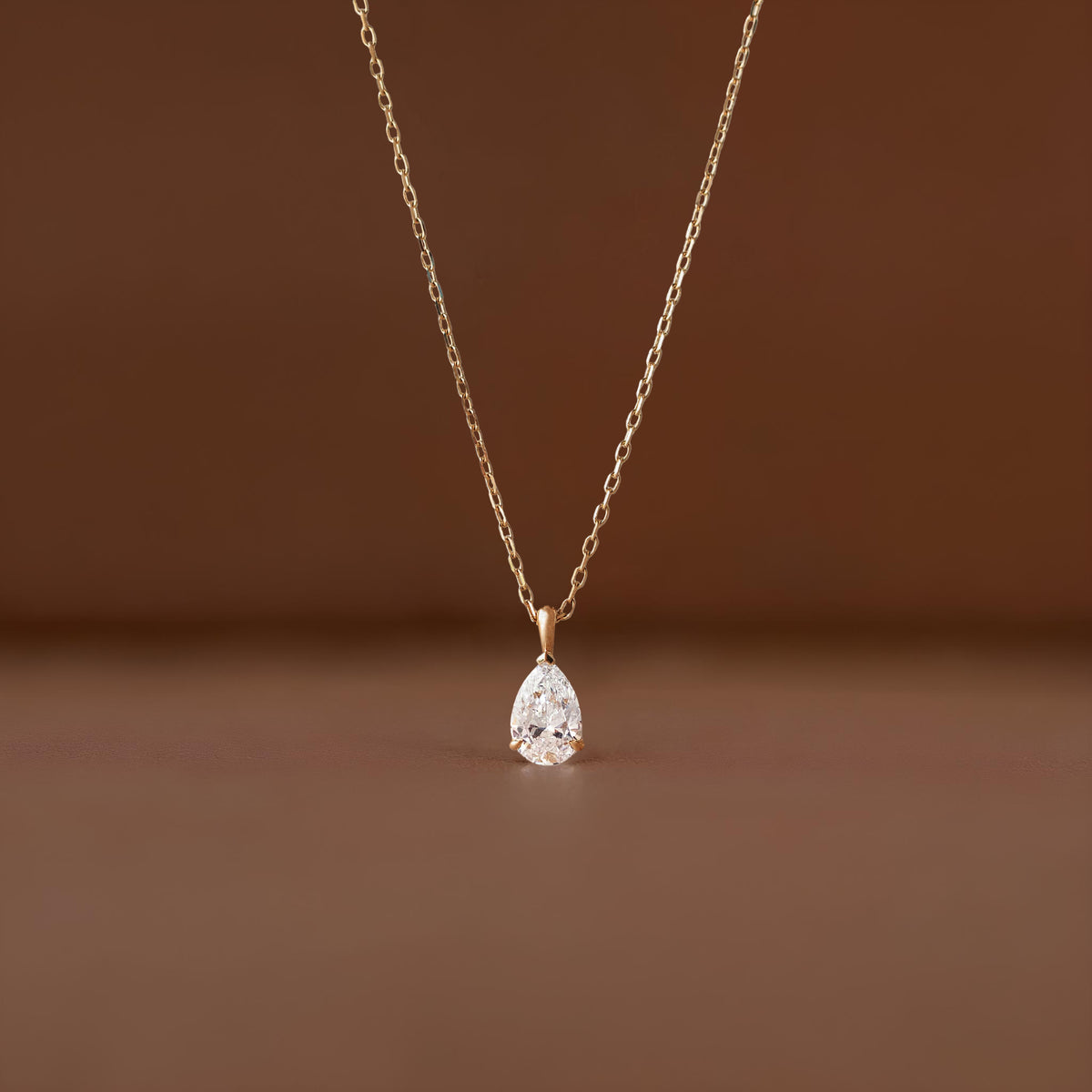A close-up shot of a delicate Gold necklace featuring a brilliant, pear-shaped diamond pendant suspended from a fine link chain against a soft, neutral brown-colored background.