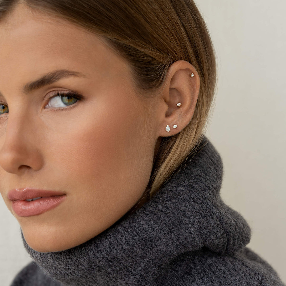 Close-up of a woman in a gray sweater, showing Gold multiple stud earrings on her ear. She gazes slightly to the side with a calm expression.