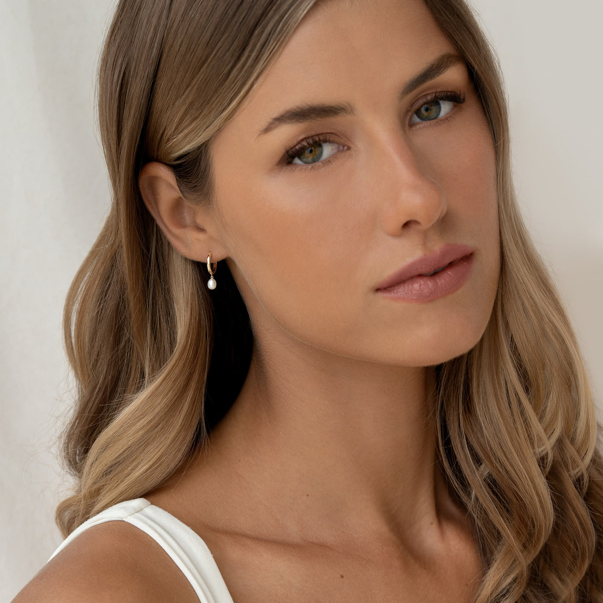 A model in a white top wears stunning 14k gold pearl drop huggie earrings, emphasizing a fashionable ensemble.