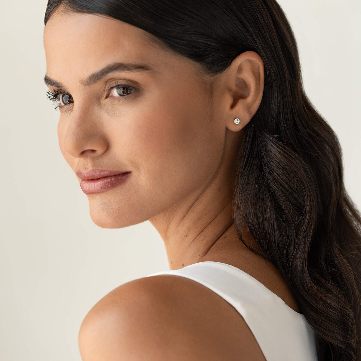 Three-quarter view of a woman wearing a small White Gold round diamond stud earring, featuring a brilliant-cut stone in a simple setting, styled with sleek dark hair and a neutral background.