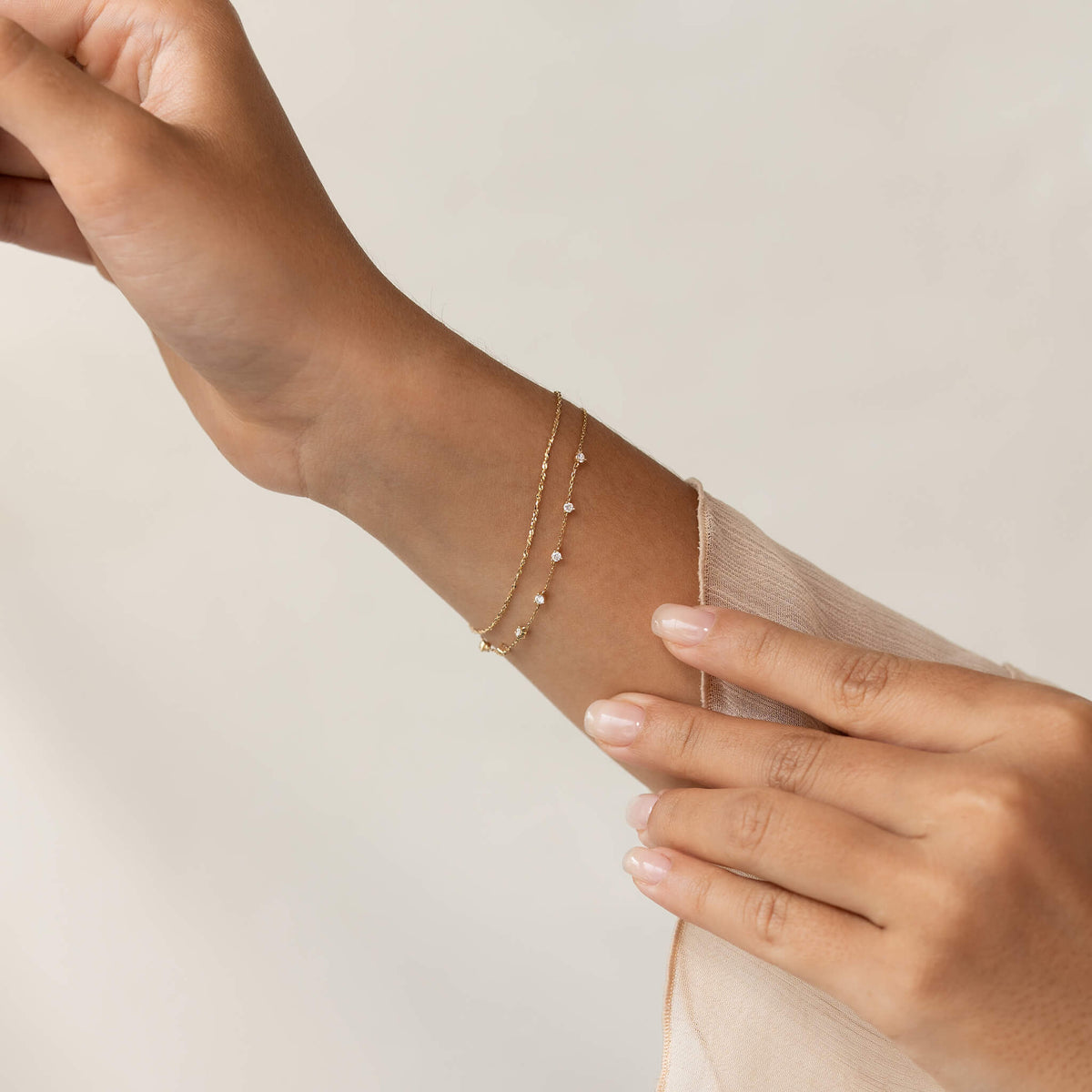 A hand gently touches a wrist adorned with delicate Gold bracelets featuring small gemstones, set against a soft, neutral background. The mood is elegant and serene.