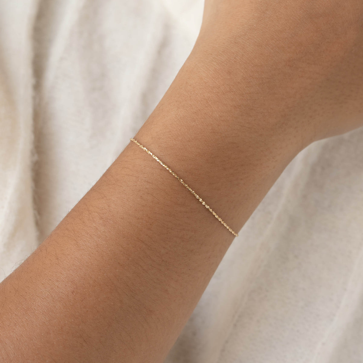 A close-up of a wrist wearing a delicate Gold chain bracelet against a soft, light-colored fabric background, conveying simplicity and elegance.