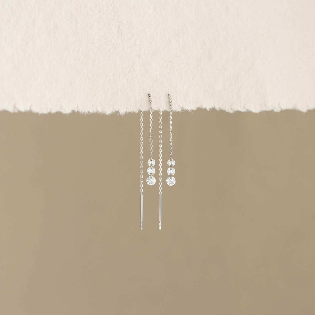 White Gold dangling earrings with sparkling beads hang against a torn paper edge and soft beige background, conveying elegance and simplicity.