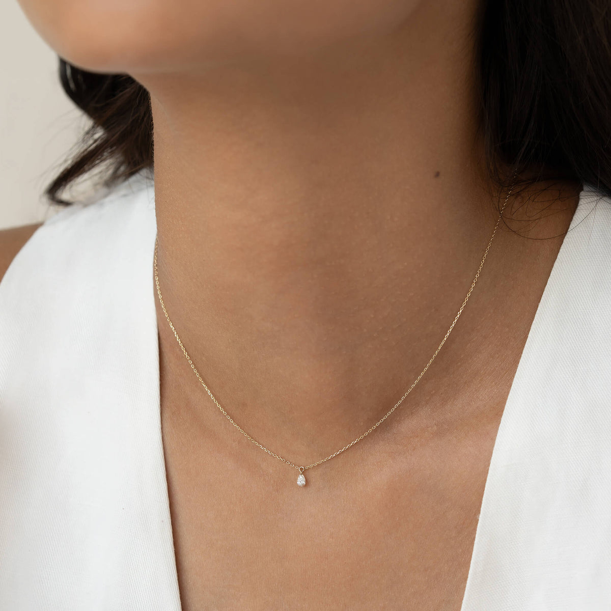 Close-up of a woman's neck wearing a delicate Gold necklace with a teardrop pendant. The background is neutral, evoking simplicity and elegance.
