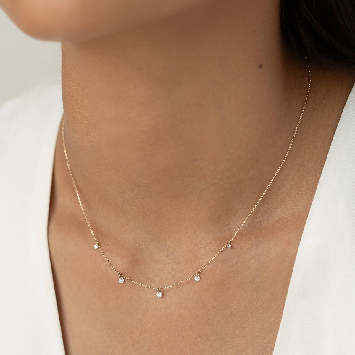 A close-up of a delicate Gold necklace with small diamond pendants adorning a woman's neck, set against a soft, neutral background.