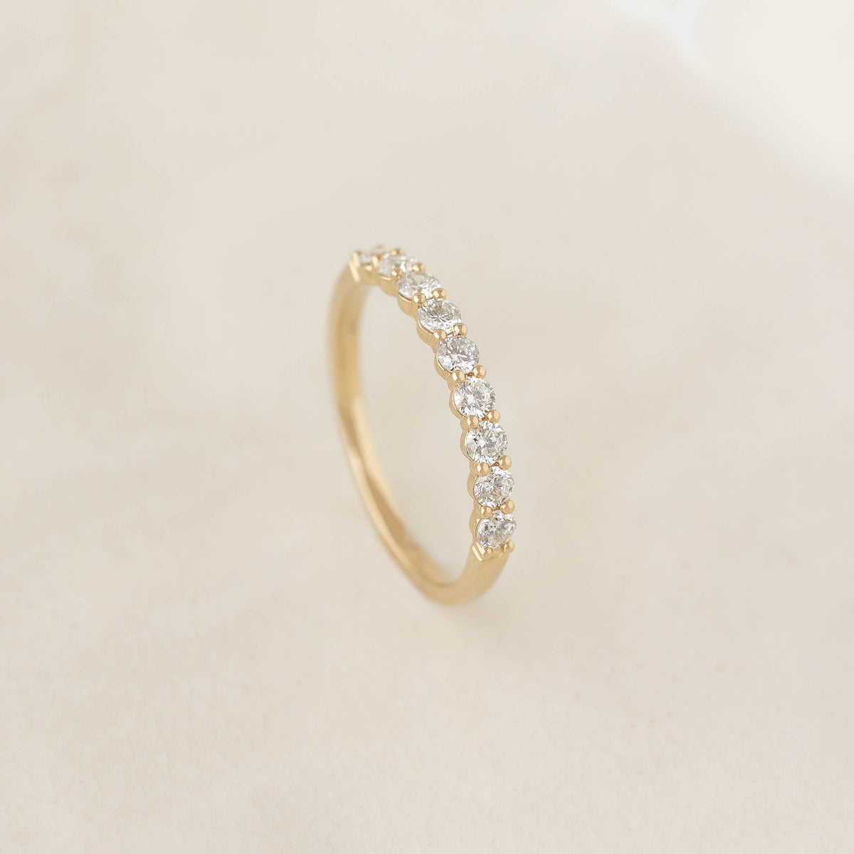 A delicate Gold ring with a row of sparkling white diamonds is showcased on a soft, beige background, conveying elegance and simplicity.