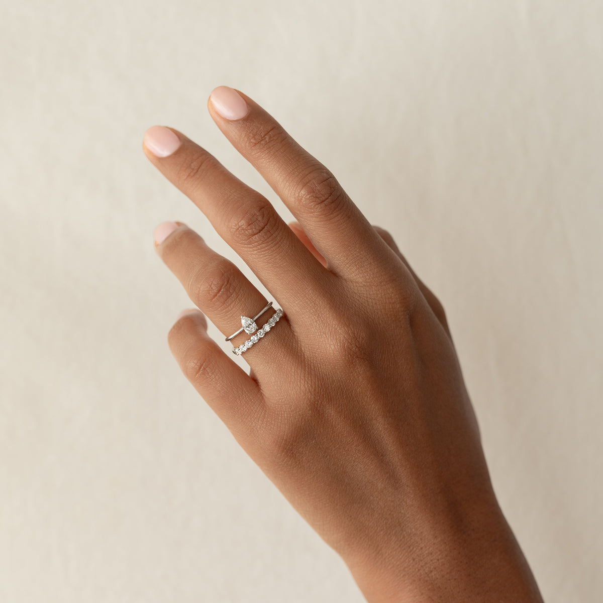 Hand with light pink nails wearing two White Gold rings, one with a pear-shaped diamond and one with a band of round diamonds.
